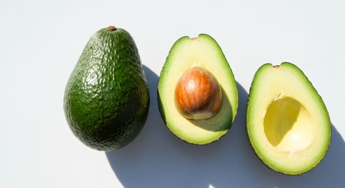 Three avocados — whole, halved showing pit, and empty half — on white surface